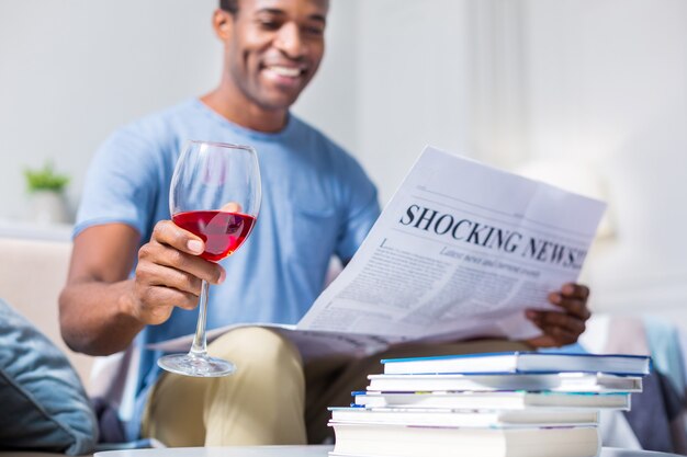 Selektiver Fokus eines Glases mit Rotwein in den Händen eines fröhlichen netten positiven Mannes beim Lesen einer Zeitung