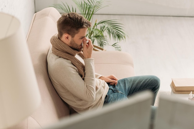 Selektiver Fokus eines kranken Mannes, der auf dem Sofa sitzt und in eine Serviette niest