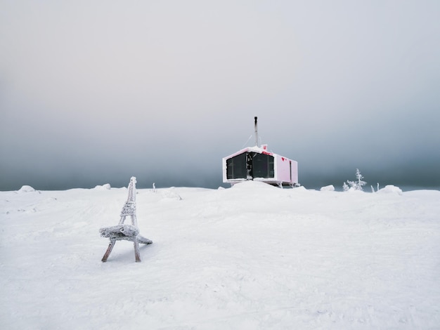 Selektiver Fokus Holzbank vor dem Hintergrund eines einsamen roten Gästehauses an einem schneebedeckten Hang im Winter unter dramatischem Himmel Doppelhaus auf dem Berg Volodyanaya Kandalaksha in Russland