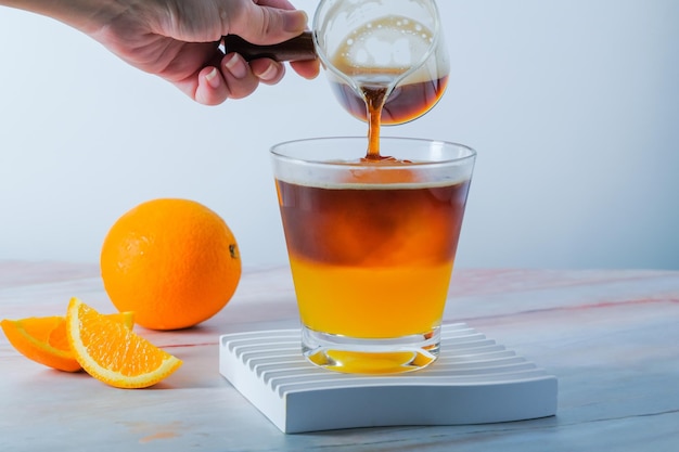 Selektiver Fokus kalt Iced Glas Espresso mit Orangensaft und frisch geschnittener Orange auf Holzplatte in weißem Hintergrund Sommercocktail Kalt gebrühter Kaffee oder schwarzer Tee Gießen kaltes Getränk