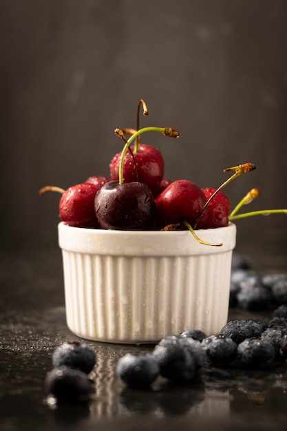 Selektiver Fokus, reife Kirschen und blaue Blaubeeren mit Wassertropfen in einer weißen Keramikschale, auf einem dunklen Hintergrund, vertikale Position