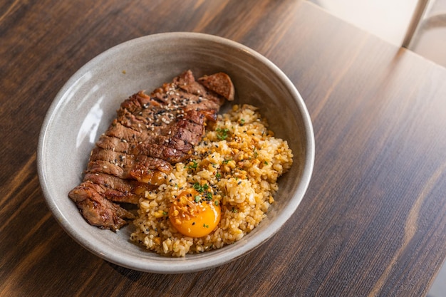 Selektiver Fokus Rinderfett Knoblauch gebratener Reis mit Rindersteak und Belag mit eingelegtem Eigelb in einer Schüssel auf Holztisch Gegrilltes Fleisch Thai ist Menü ist Fusion Food