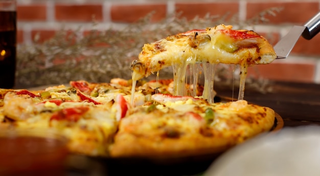 Selektiver Fokus und Nahaufnahme auf einem Stück hausgemachter Meeresfrüchtepizza mit doppeltem Käse auf Spachtel oder Schaufel mit unscharfem Hintergrund aus Holztisch, kaltem Getränk und Ziegelwand. Lebensmittelkonzept.