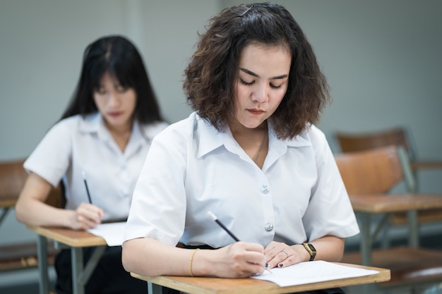 Selektiver Fokus von College-Studenten im Teenageralter sitzt auf dem Lehrstuhl, schreibt auf dem Prüfungspapier Antwortbogen und nimmt den Abschlussprüfungsraum oder das Klassenzimmer auf. Universitätsstudenten in Uniform im Klassenzimmer.