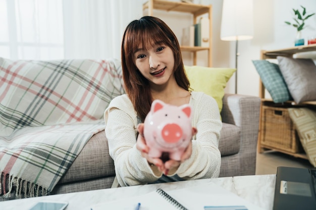 Selektives Fokusporträt eines süßen asiatischen Mädchens, das in die Kamera lächelt, zeigt ein Sparschwein in Händen mit ausgestreckten Armen in einem gemütlichen Wohnzimmer zu Hause.