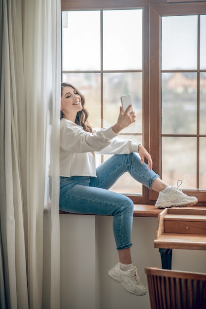 Foto selfie. bastante joven mujer de pelo largo con una blusa blanca sentada en la ventana y haciendo selfie