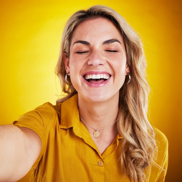 Selfie cara y sonrisa con una mujer que se ríe sobre un fondo amarillo en el estudio por diversión o humor Ojo cerrado cómico y divertido con una atractiva joven modelo que disfruta de la comedia siendo tonta o tonta