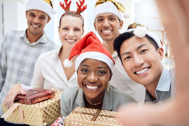 Selfie Christmas y empleados con regalo en una celebración de fiesta y sonrisa en la oficina juntos en el trabajo Retrato de jóvenes felices y equipo de trabajadores de negocios con regalo y foto en evento corporativo