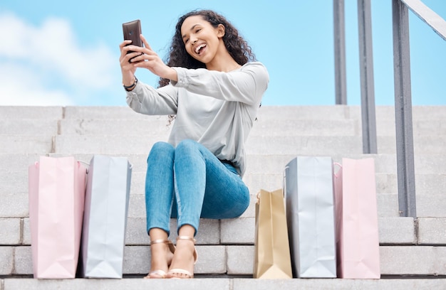 Selfie de compras y una mujer influyente y bloguera sentada afuera con bolsas después de una juerga de gastos mientras estaba en las escaleras de la ciudad Consumidor feliz o cliente transmitiendo en vivo oferta de promoción y venta de moda