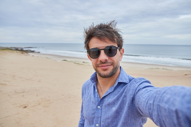 Selfie de homem branco sorrindo na praia com óculos escuros e camisa azul clara.