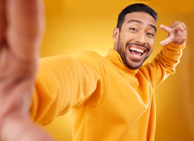 Selfie de sinal de paz e retrato feliz de um homem no estúdio com um emoji de mão e um sorriso Modelo asiático masculino animado em um fundo amarelo com um ícone positivo para foto de perfil de mídia social