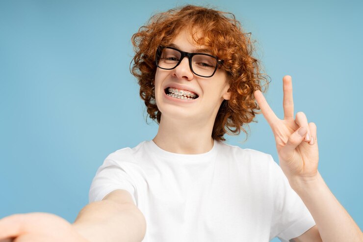 Foto selfie de um adolescente sorridente com cabelo vermelho encaracolado e aparelhos ortopédicos usando óculos e fazendo sinal de paz