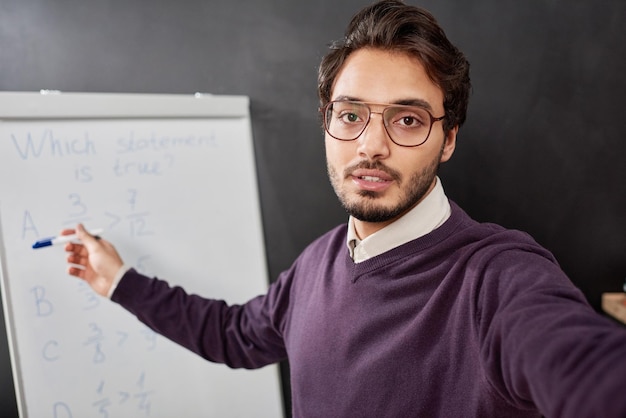 Selfie eines selbstbewussten jungen Mischlingslehrers mit Brille und violettem Pullover, der auf Whiteboard zeigt