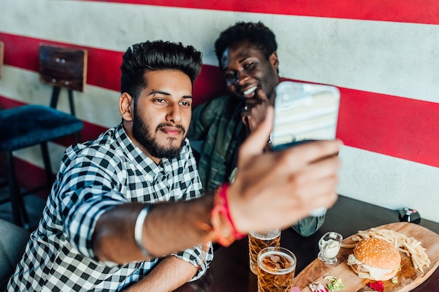 Selfie engraçado. Retrato de amigos tirando foto com o smartphone enquanto está sentado à mesa com cerveja e petiscos.