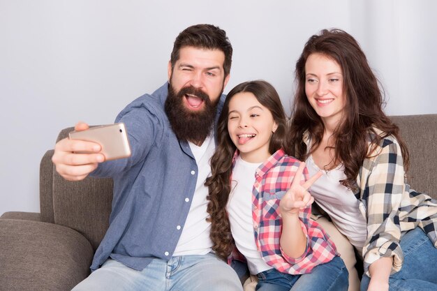 Selfie Familia feliz en casa Teléfono inteligente moderno Niña usa teléfono inteligente con madre y padre hombre barbudo y mujer con hija Selfie mania Familia hace selfie en teléfono inteligente Capturando diversión