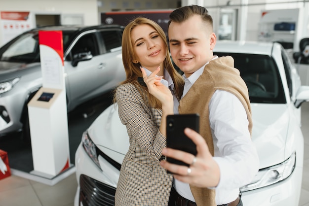 Selfie familiar en concesionario. Feliz pareja joven elige y compra un coche nuevo para la familia.