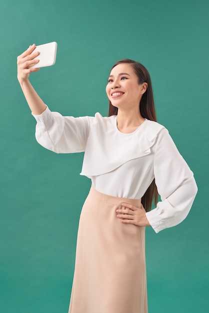 Selfie! Fröhliche junge asiatische Frau, die Handy hält und Selfie mit ihrem Smartphone macht, während sie vor grünem Hintergrund steht