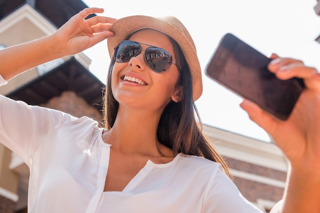 Selfie für meinen Freund. Niedrige Winkelsicht der schönen jungen funky Frau, die ihren Hut justiert und selfie macht, während sie im Freien steht