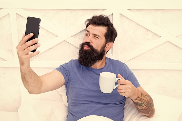 Selfie kiss Hombre barbudo se toma selfie en la cama Hipster sonríe a la cámara selfie en el teléfono móvil Disfrutando de una sesión de selfie Rutina de café matutino Vida moderna Nueva tecnología Red social