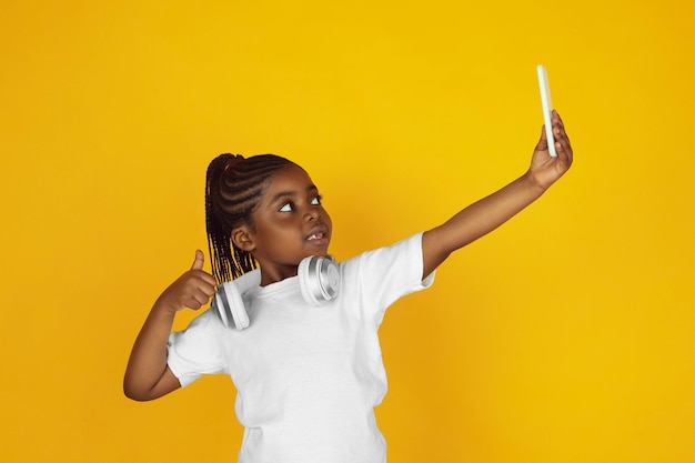 Selfie machen, Musik hören. Porträt des kleinen afroamerikanischen Mädchens auf gelbem Studiohintergrund. Fröhliches Kind. Konzept der menschlichen Emotionen, Gesichtsausdruck, Verkauf, Anzeige. Exemplar. Sieht süß aus.
