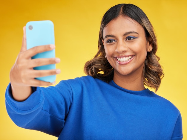 Selfie mídia social e sorriso com uma mulher em um fundo amarelo no estúdio posando para uma foto de perfil Fotografia de rosto e um jovem influenciador feliz dentro de casa para atualizar um status em sua linha do tempo