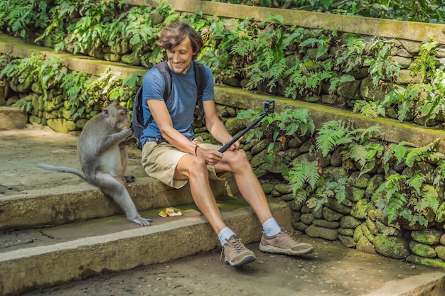 Selfie con monos. El joven usa un palo selfie para tomar una foto o un video blog con un lindo mono divertido. Selfie de viaje con vida silvestre en Bali.