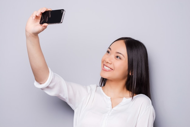 Selfie! mulher asiática jovem alegre segurando um telefone celular e fazendo selfie com seu telefone inteligente em pé contra um fundo cinza