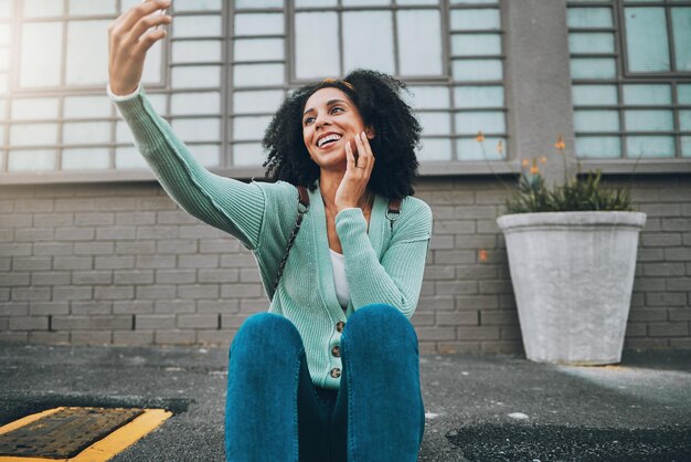 Selfie mulher negra e smartphone na cidade feliz e sorriso na transmissão ao ar livre ao vivo e na calçada Jovem mulher e telefone sendo alegre na moda e nervoso na calçada para a felicidade