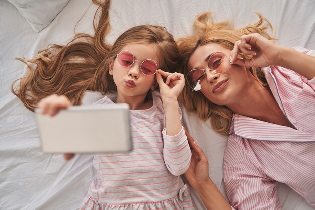 Selfie perfeito. Vista superior da bela jovem mãe e sua linda filha tirando uma selfie com o telefone inteligente e sorrindo enquanto estavam deitada na cama em casa