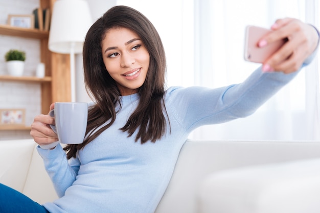 Selfie por dia. mulher alegre e alegre tomando café enquanto usa o smartphone