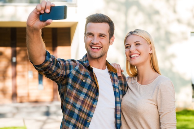 Selfie para su álbum familiar. Pareja joven alegre que se une entre sí mientras hace selfie por teléfono inteligente mientras está de pie contra su nueva casa