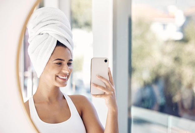 Selfie de teléfono de retrato y toalla de mujer y baño para el cuidado de la piel, belleza y limpieza en el reflejo del espejo Cara de niña y sonrisa para la foto después del tratamiento facial, aseo e higiene con una sonrisa