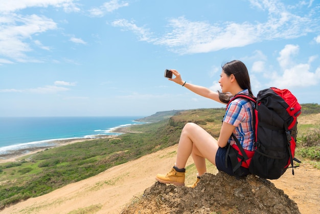 selfie turista mochilero sentado en la montaña feliz con hermosos paisajes tomando fotos a través de un teléfono inteligente móvil con fondo de cielo oceánico.