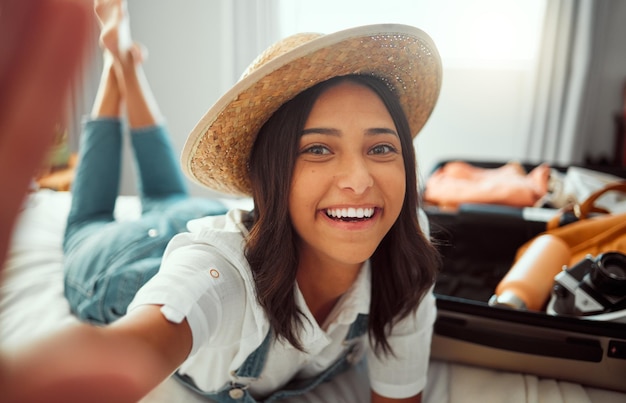 Selfie de viaje facial y mujer empacando maleta preparándose para vacaciones o vacaciones Retrato relajado y mujer feliz tomando fotos con sombrero mientras carga equipaje en la cama en preparación para el viaje