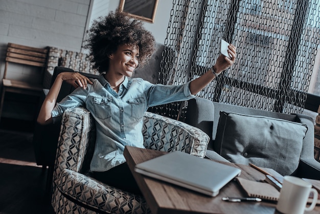 Selfie Zeit. Schöne junge Afrikanerin, die selfie mit ihrem Smartphone macht, während sie im Café sitzt sitting