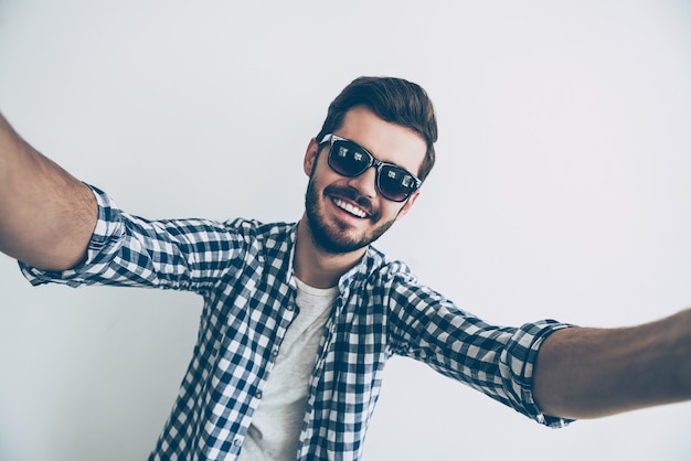 Selfie Zeit! Studioaufnahme eines gutaussehenden jungen Mannes mit Sonnenbrille, der Selfie macht und lächelt