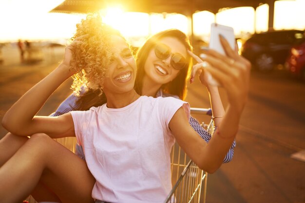 Selfie-Zeit Zwei schöne Mädchen, die Spaß auf Einkaufswagen haben Junge Leute, die auf Einkaufswagen laufen