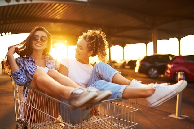 Selfie-Zeit Zwei schöne Mädchen, die Spaß auf Einkaufswagen haben Junge Leute, die auf Einkaufswagen laufen