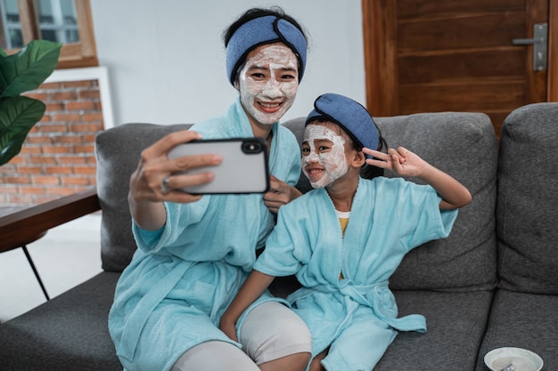 Selfie zusammen mit Handy der asiatischen Mutter und des kleinen Mädchens machen Gesichtsmaske, wenn Sie zu Hause entspannen