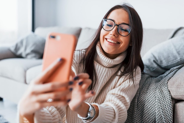 Selfies machen. Junge schöne Frau mit Brille sitzt allein zu Hause mit Telefon in den Händen.