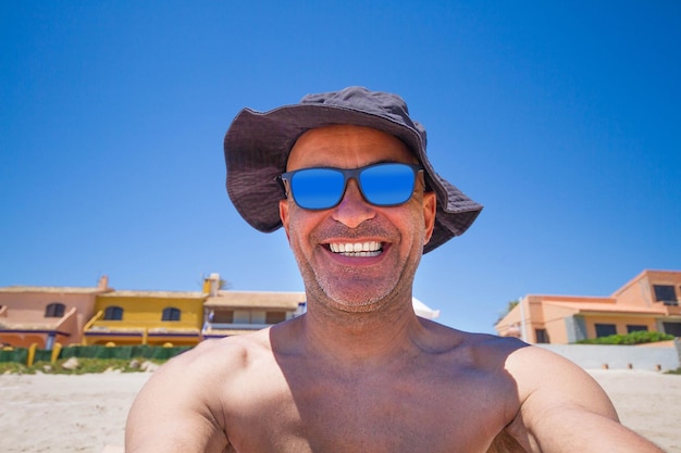 Foto selfy de um homem de meia idade em um chapéu e óculos de sol sorrindo alegremente na praia