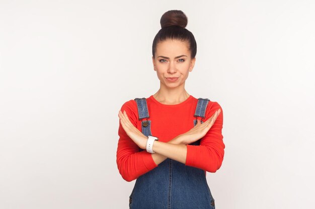 Sem acesso Retrato de menina séria estrita com coque de cabelo em macacão jeans mostrando sinal de pare com as mãos cruzadas, rejeitando a expressão de aviso de oferta tiro de estúdio interno isolado no fundo branco