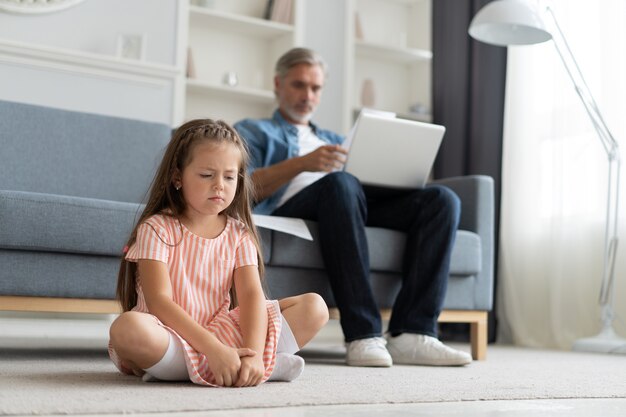 Sem tempo para a criança. Pai grisalho ocupado com o laptop, trabalhando online em casa, filha triste entediada ofendida sentada perto.