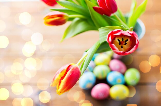 Foto semana santa, vacaciones, tradición y concepto de objeto: cierre de huevos de pascua de colores y flores de tulipán en un cubo sobre una mesa de madera durante las luces navideñas