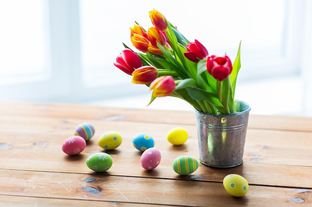 semana santa, vacaciones, tradición y concepto de objeto: cierre de huevos de pascua de colores y flores de tulipán en un cubo sobre una mesa de madera