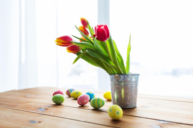 semana santa, vacaciones, tradición y concepto de objeto: cierre de huevos de pascua de colores y flores de tulipán en un cubo sobre una mesa de madera