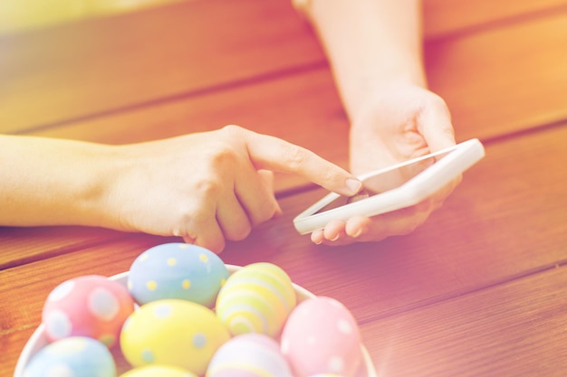 Semana Santa, vacaciones, tradición, tecnología y concepto de personas: cierre de manos de mujer con huevos de Pascua de colores en el plato y el teléfono inteligente