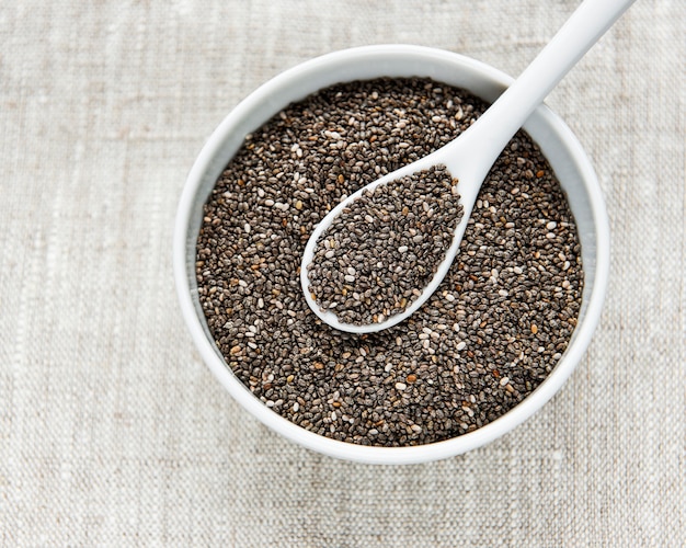 Sementes de Chia em uma tigela branca e uma colher em fundo de matéria têxtil.