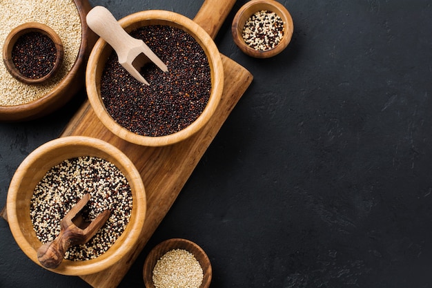 Sementes de grãos de quinua branca, preta e mista em tigelas de bambu em uma mesa de pedra preta. Postura plana com espaço de cópia.