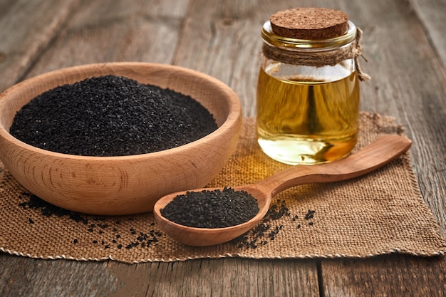 Semillas de comino negro y una cuchara de madera, recipiente con botella de aceite sobre una mesa de madera.